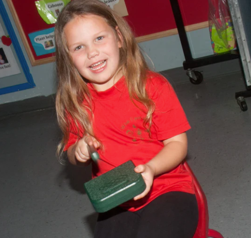 Child seated on red chair holding green tone block with diamond-patterned etching and striker, indoor setting