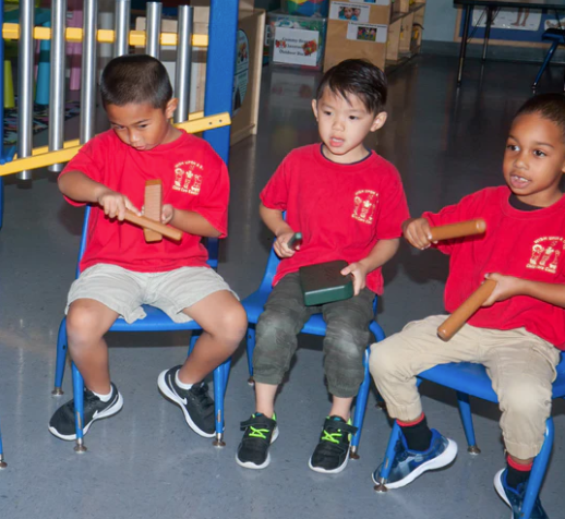 Three children playing wooden percussion instruments including guiros and rhythm blocks in a classroom setting