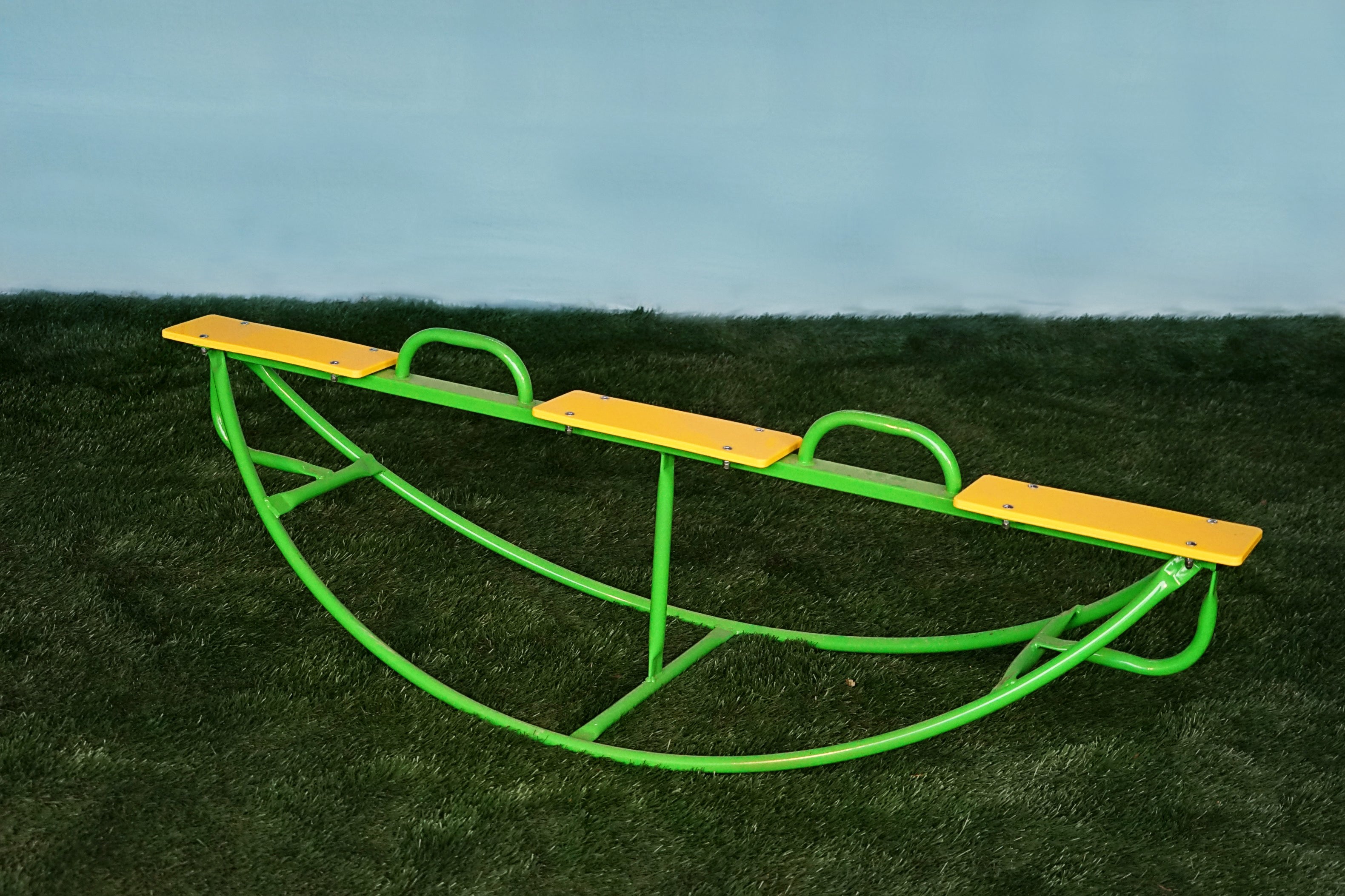 Green powder-coated steel see saw with three yellow seats and handlebars on artificial grass, designed for safe children's play.