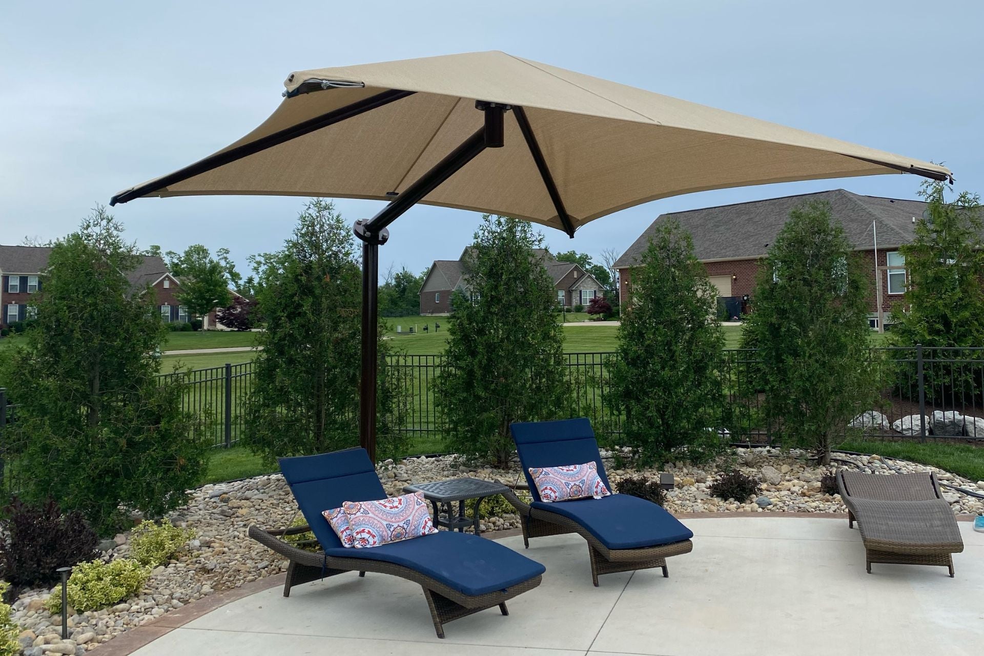 Beige pyramid cantilever shade with single dark post over two blue cushioned lounge chairs on patio