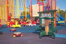 Children seated around a colorful green and brown outdoor musical drum table with hand drums on rubberized flooring