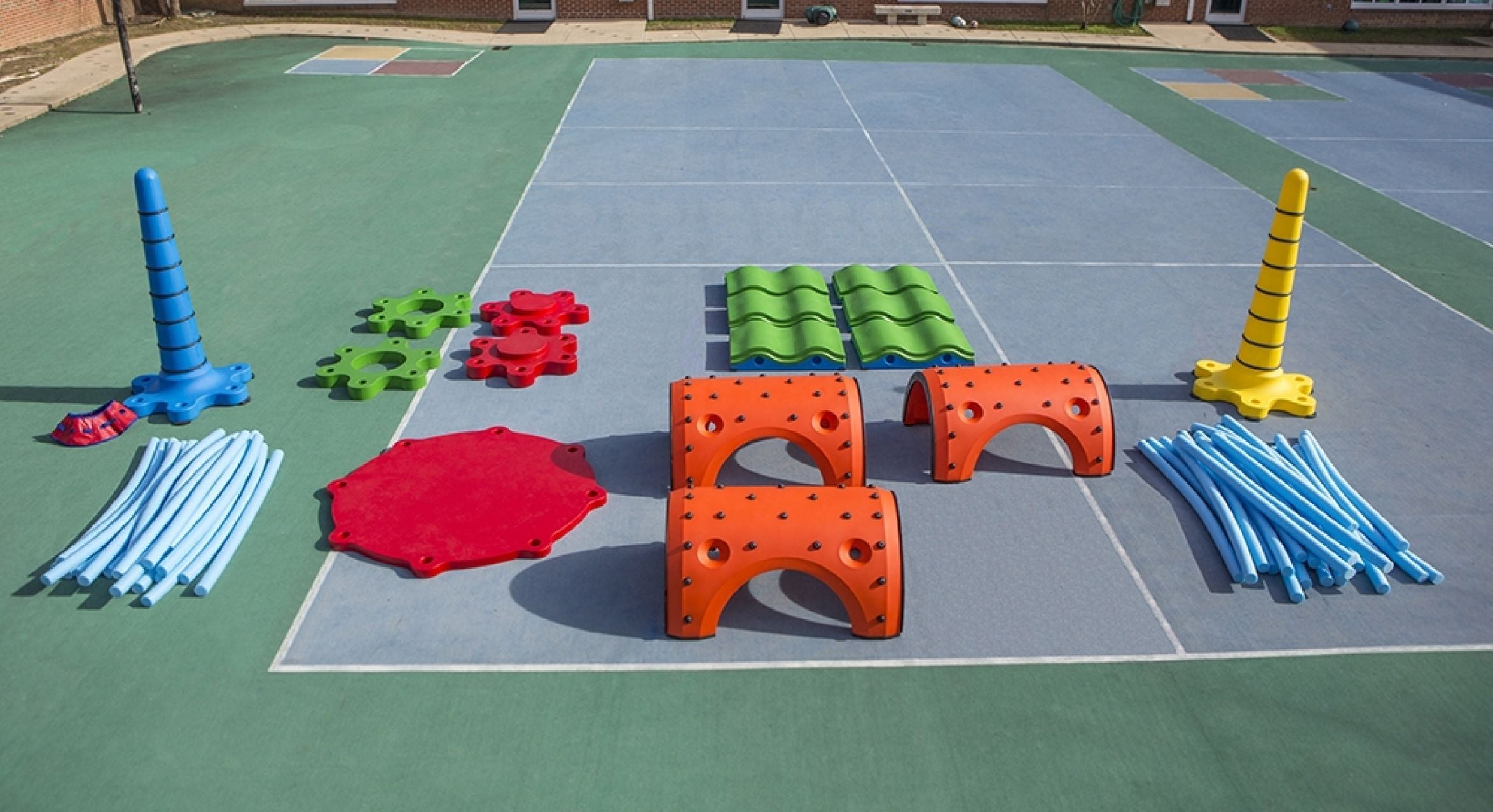 Colorful modular playground pieces including orange arches, green wave panels, blue flexible noodles, and tall cone-shaped elements on a sports court
