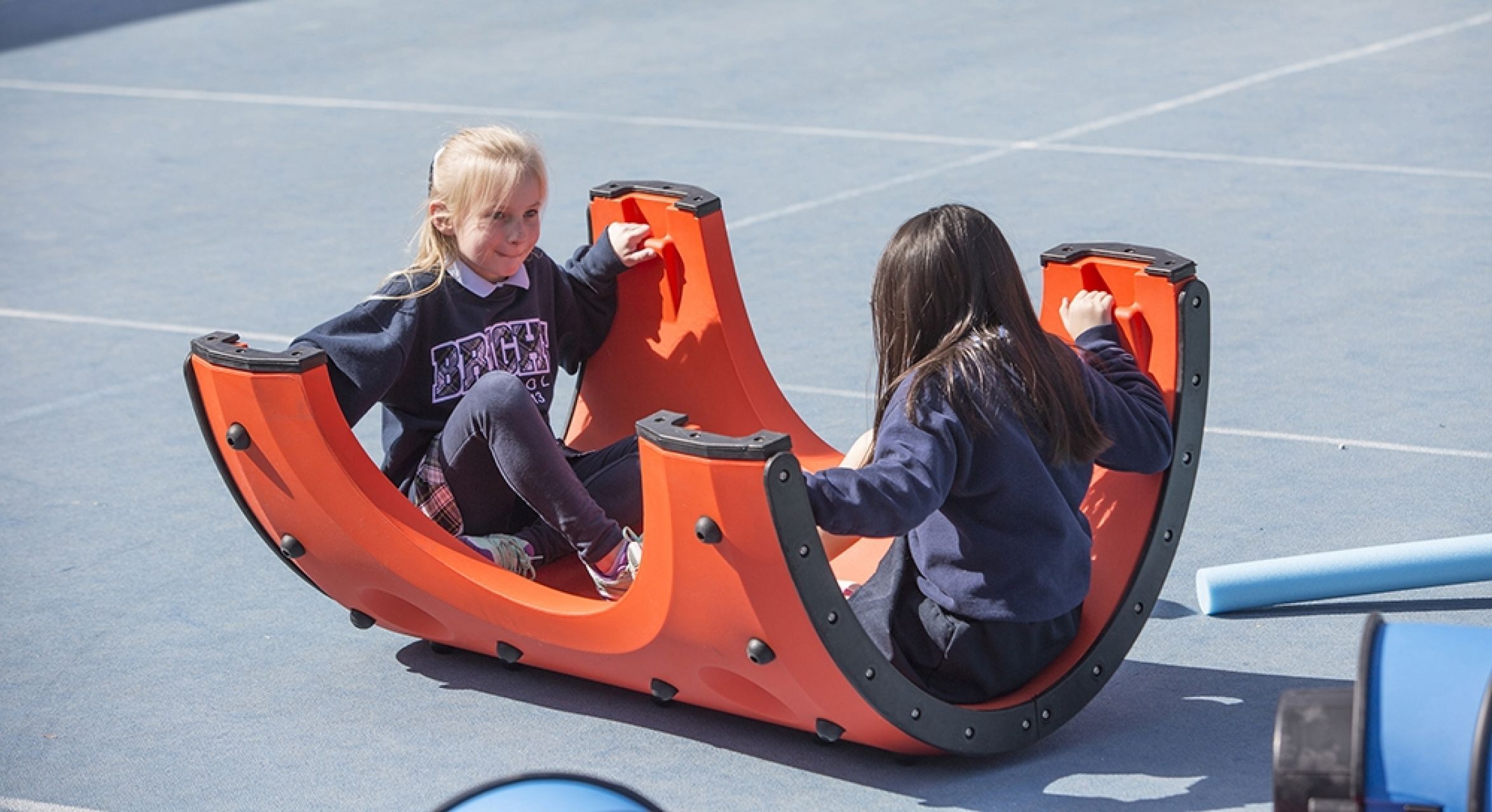 Two children seated facing each other on a curved orange interactive play structure with hand grips on a blue outdoor surface