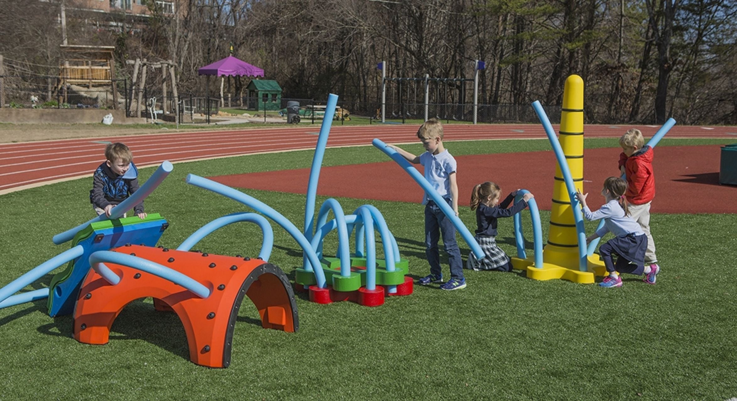 Children playing on colorful outdoor interactive playground equipment with blue flexible rods on artificial turf near a running track