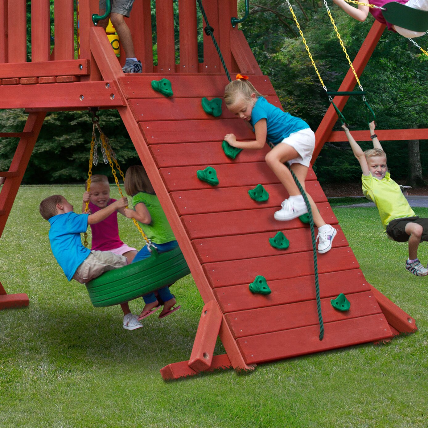 Redwood-finished wooden swing set with green climbing holds, rope ladder, tire swing, and children playing on grass