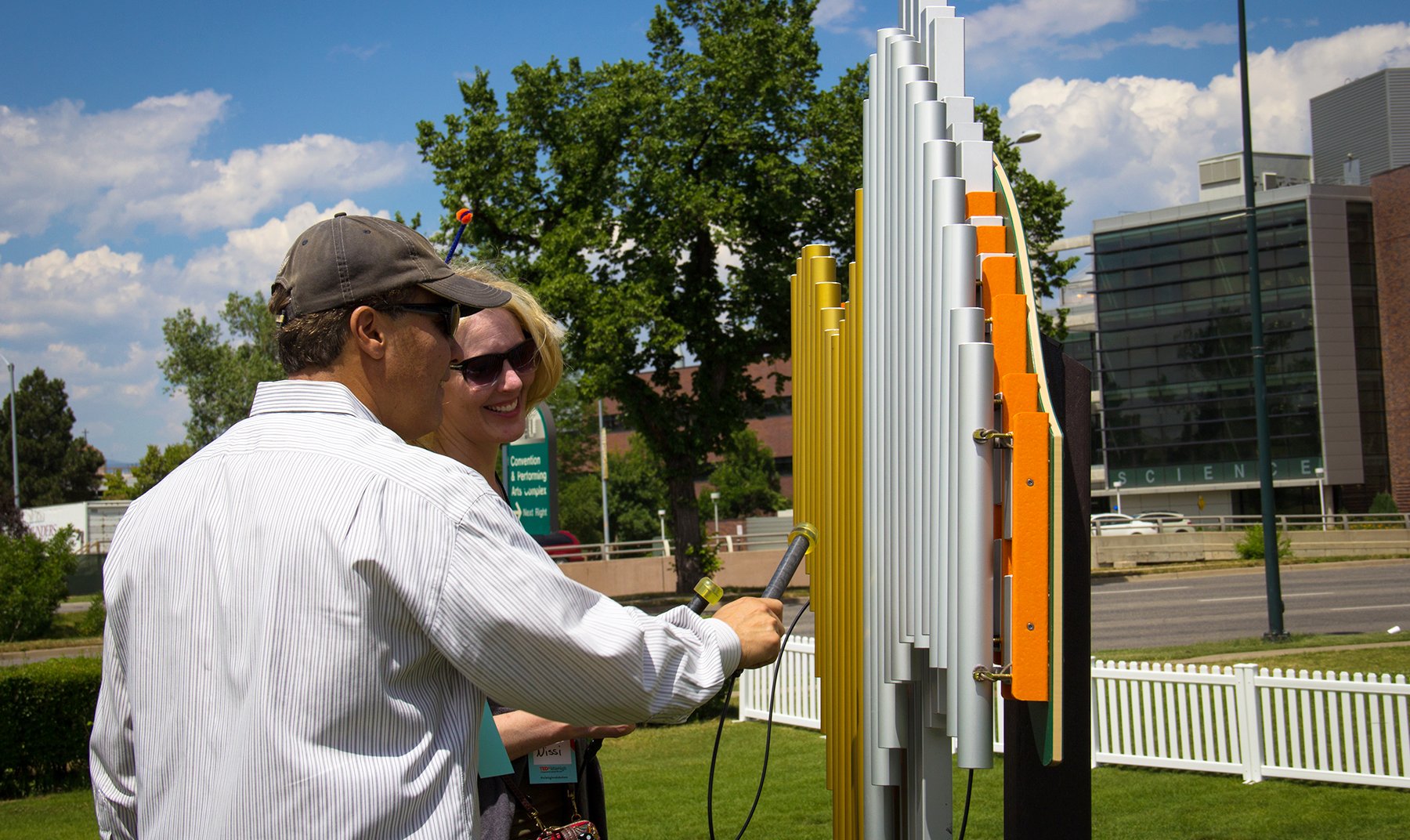 Swirl Outdoor Musical Park Instrument with 26 anodized aluminum chimes in vivid orange, played by two adults outdoors