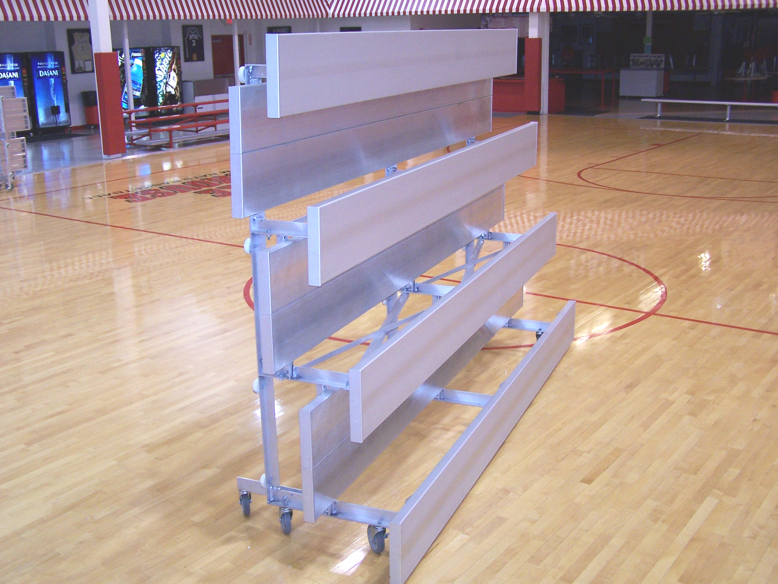 Folded aluminum low-rise bleachers with four rows on swivel casters, positioned on a gymnasium floor