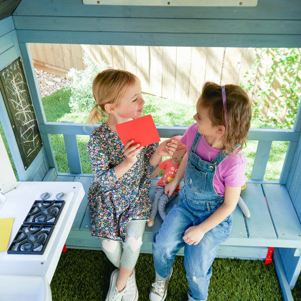 Two girls playing with dolls and cards inside a blue wooden playhouse featuring a built-in bench, toy stove, and chalkboard.