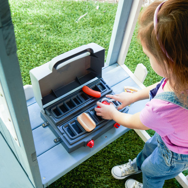 Child playing with toy grill and plastic hotdog on wooden playhouse patio with green grass outdoors