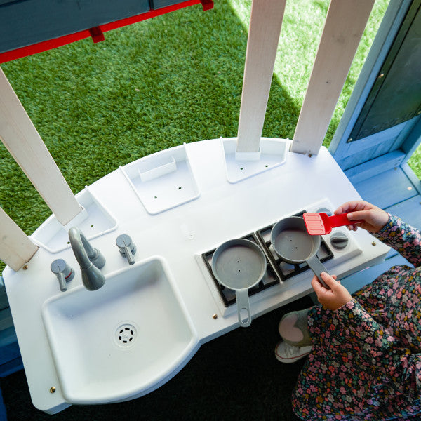 Overhead view of child playing with white plastic toy kitchen sink and stove on wooden playhouse patio with picket fence and gate