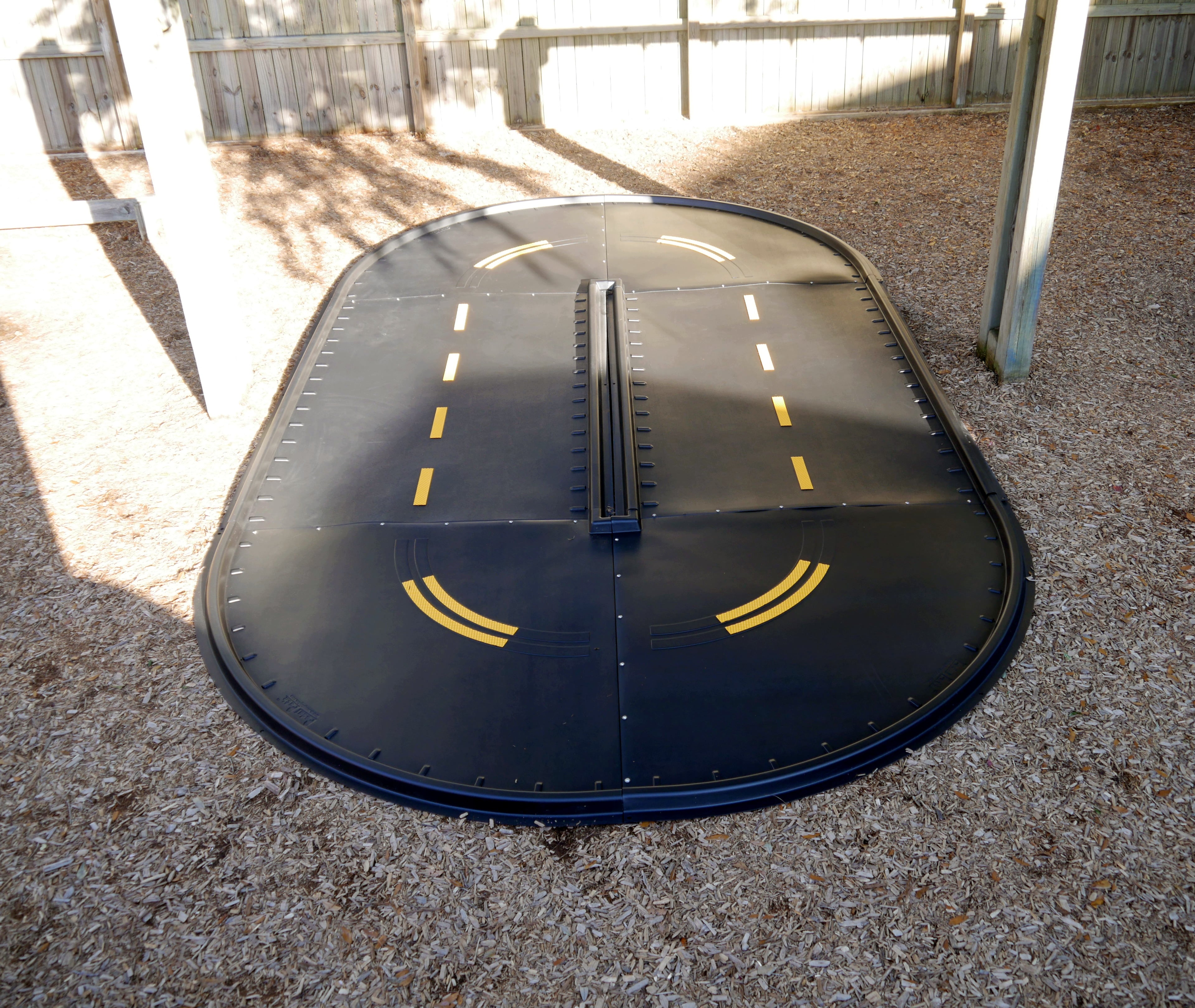 Oval-shaped black trike path with yellow lane markings and central divider on wood chip surface, designed for accessible active play.