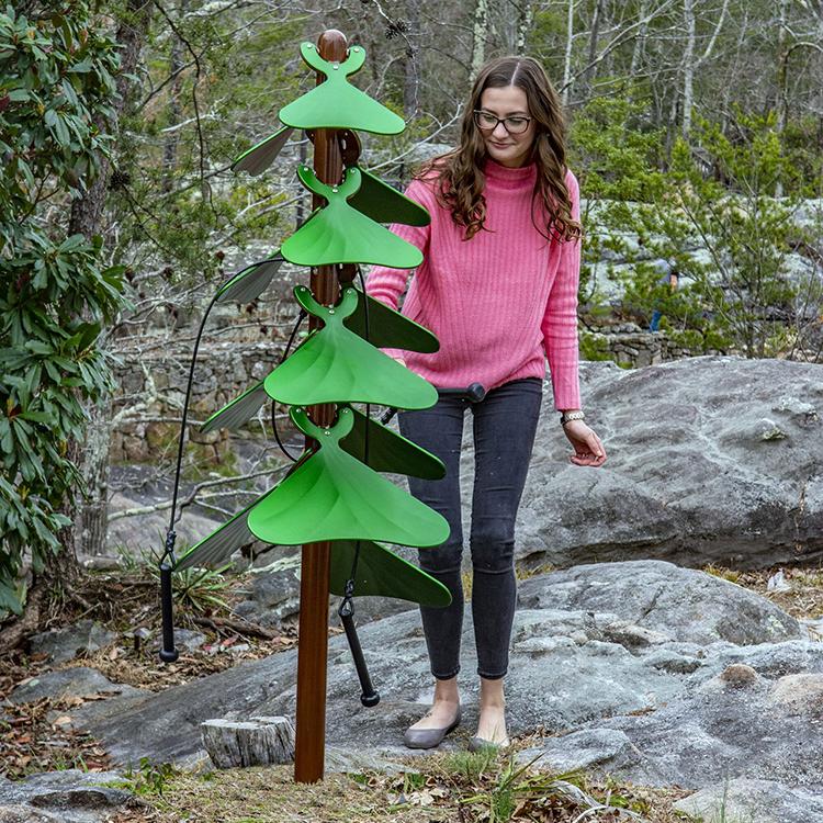 Outdoor musical instrument with green anodized aluminum leaves on brown post, played by a woman in a natural setting
