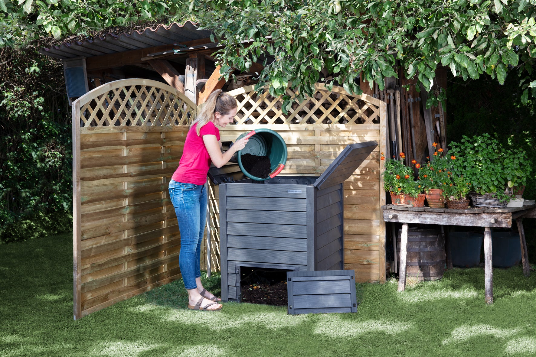 Woman adding soil to dark gray insulated compost bin with hinged lids and bottom removal doors in garden setting