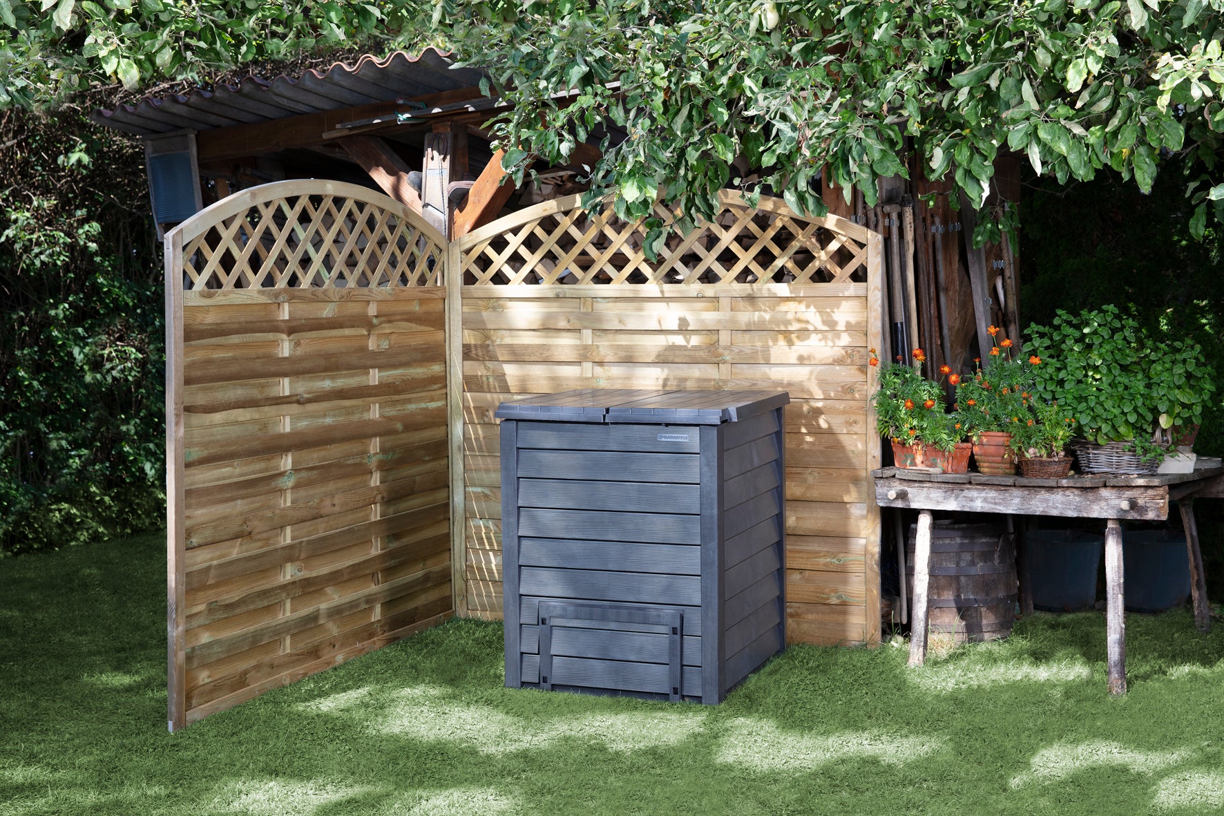 Dark gray insulated compost bin with hinged top lids, bottom removal doors, and hidden side ventilation, placed on grass near garden lattice and table with plants