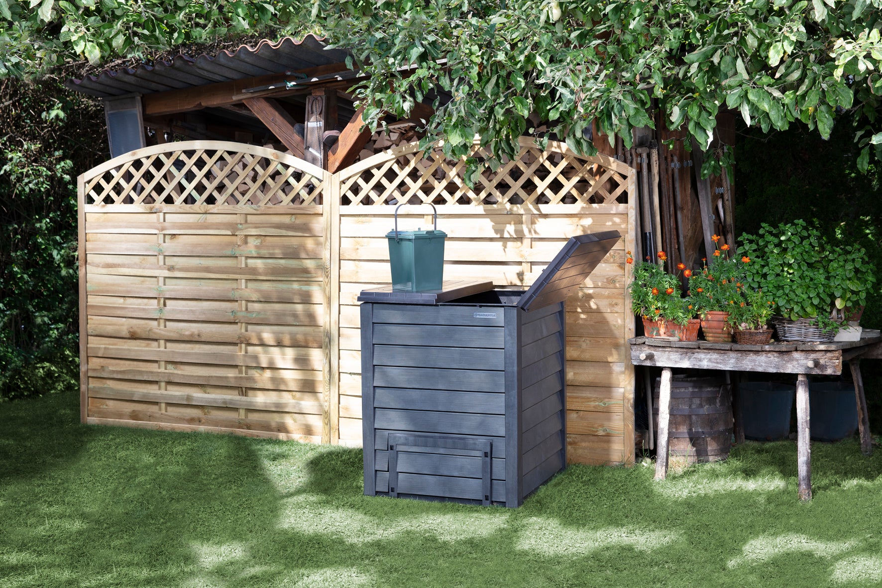 Dark gray compost bin with hinged top lids and dual bottom doors, textured like wood, on grass near garden fence and table with plants