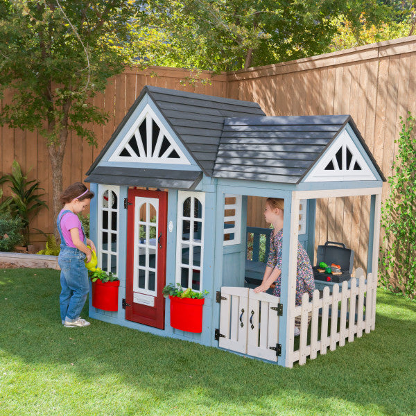 Wooden outdoor playhouse with red door, white picket fence patio, planter boxes, and two children playing nearby