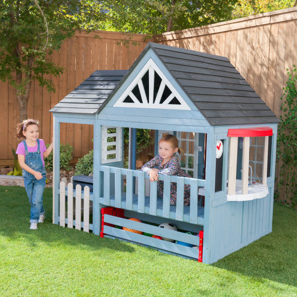 Blue wooden outdoor playhouse with black roof, white picket fence, bay window, and children playing in a backyard setting