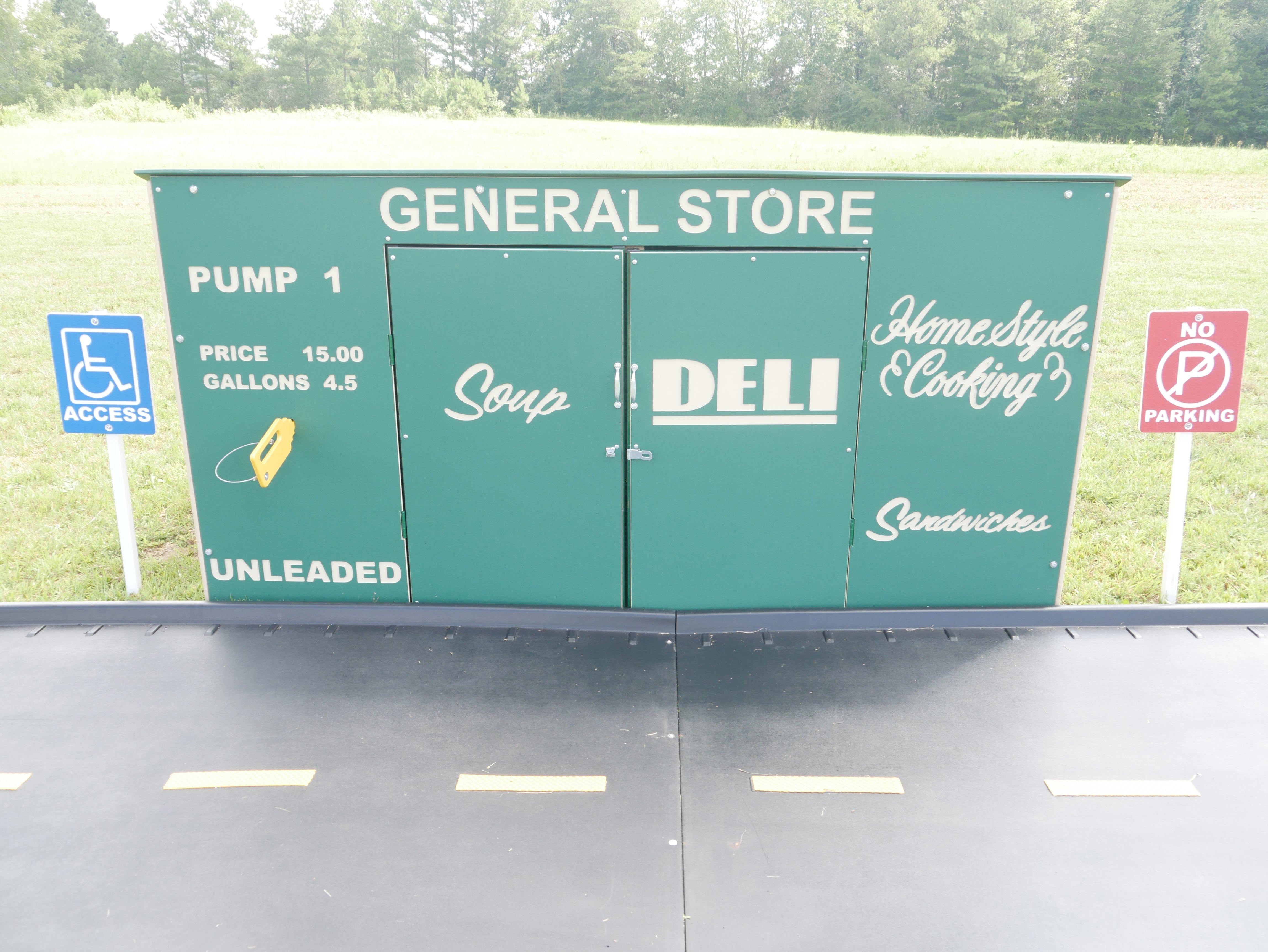 Green playhouse styled as a general store with doors, gas pump graphics, accessibility and no parking signs, durable outdoor play structure