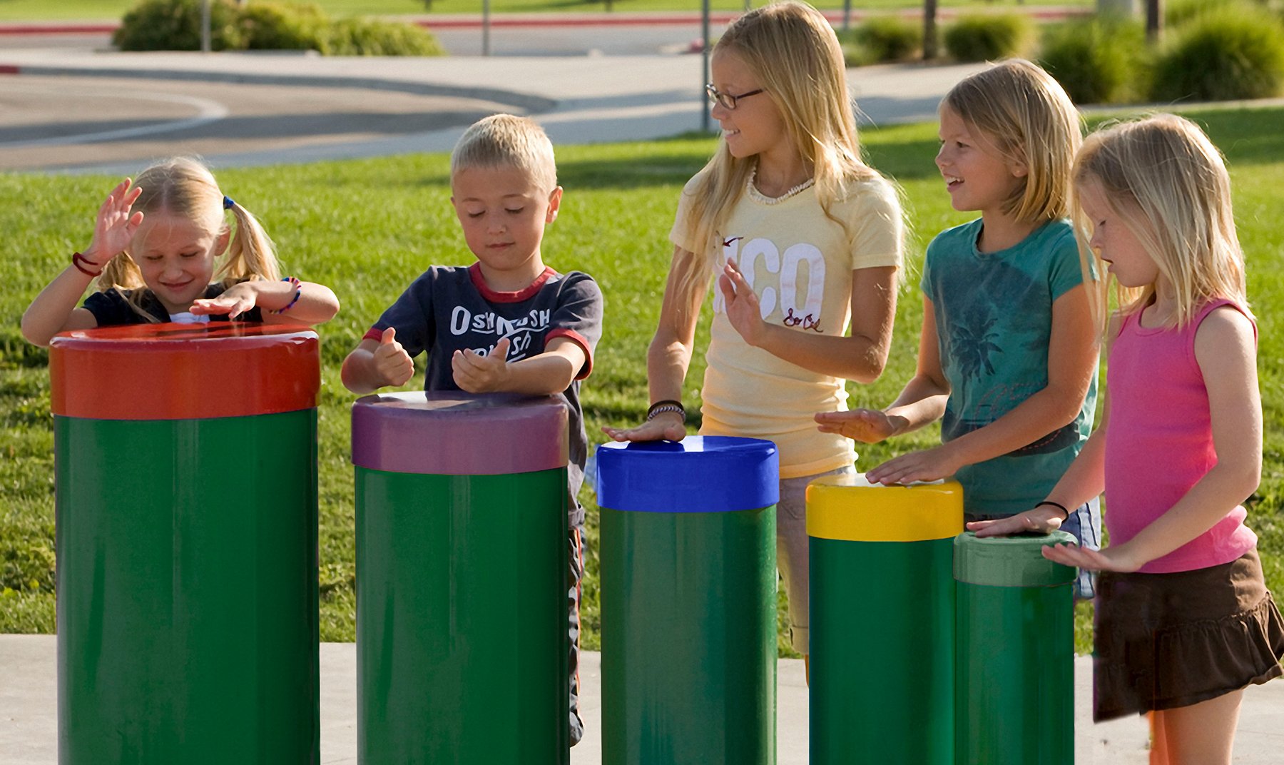 Five children playing colorful toddler-height tuned drums with vibrant tops on green bases outdoors in a park setting