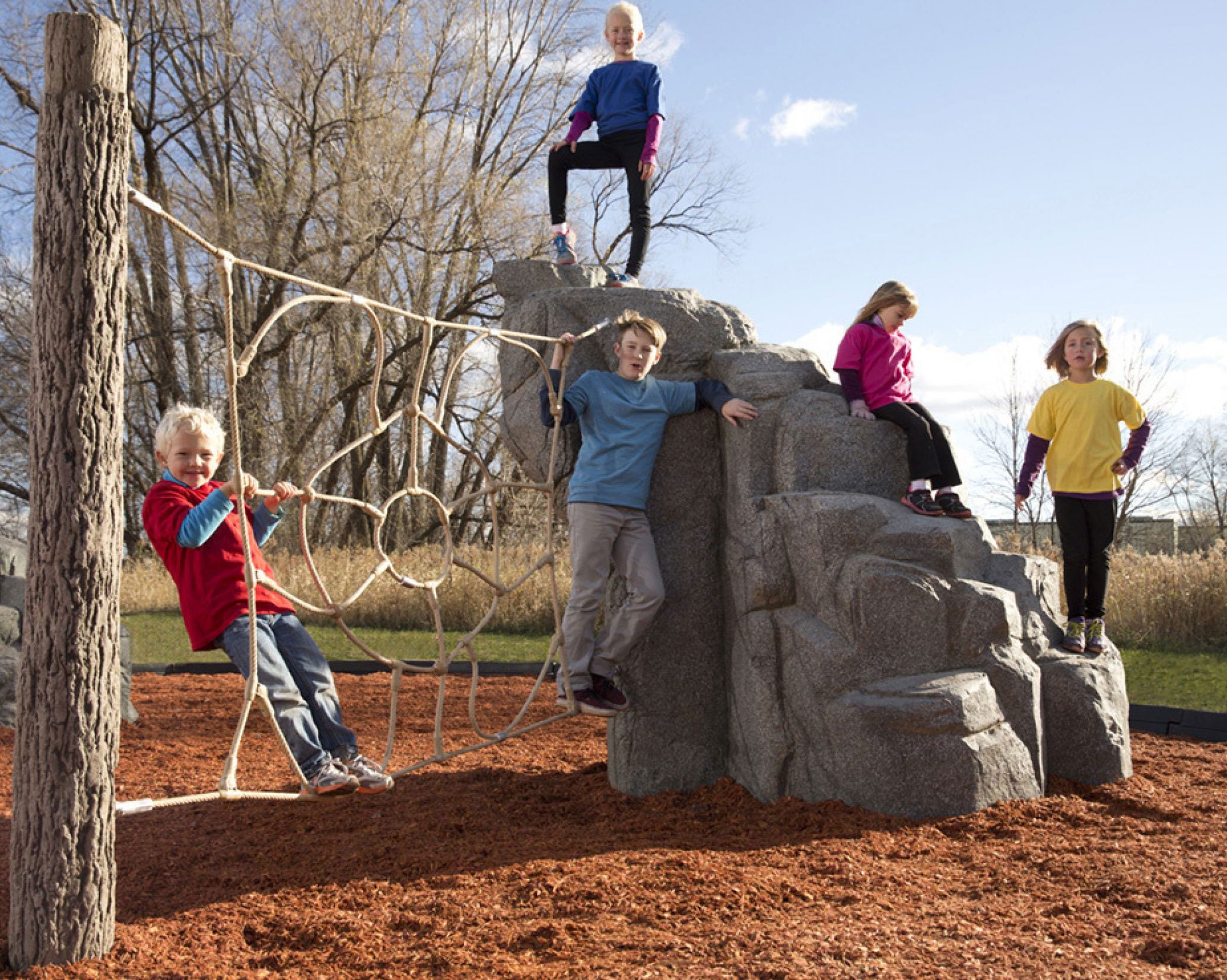 Children playing on the Nature Rocks Spider Mountain Climbing Rock, featuring a granite boulder, log post, and spider web net in an outdoor playground setting.