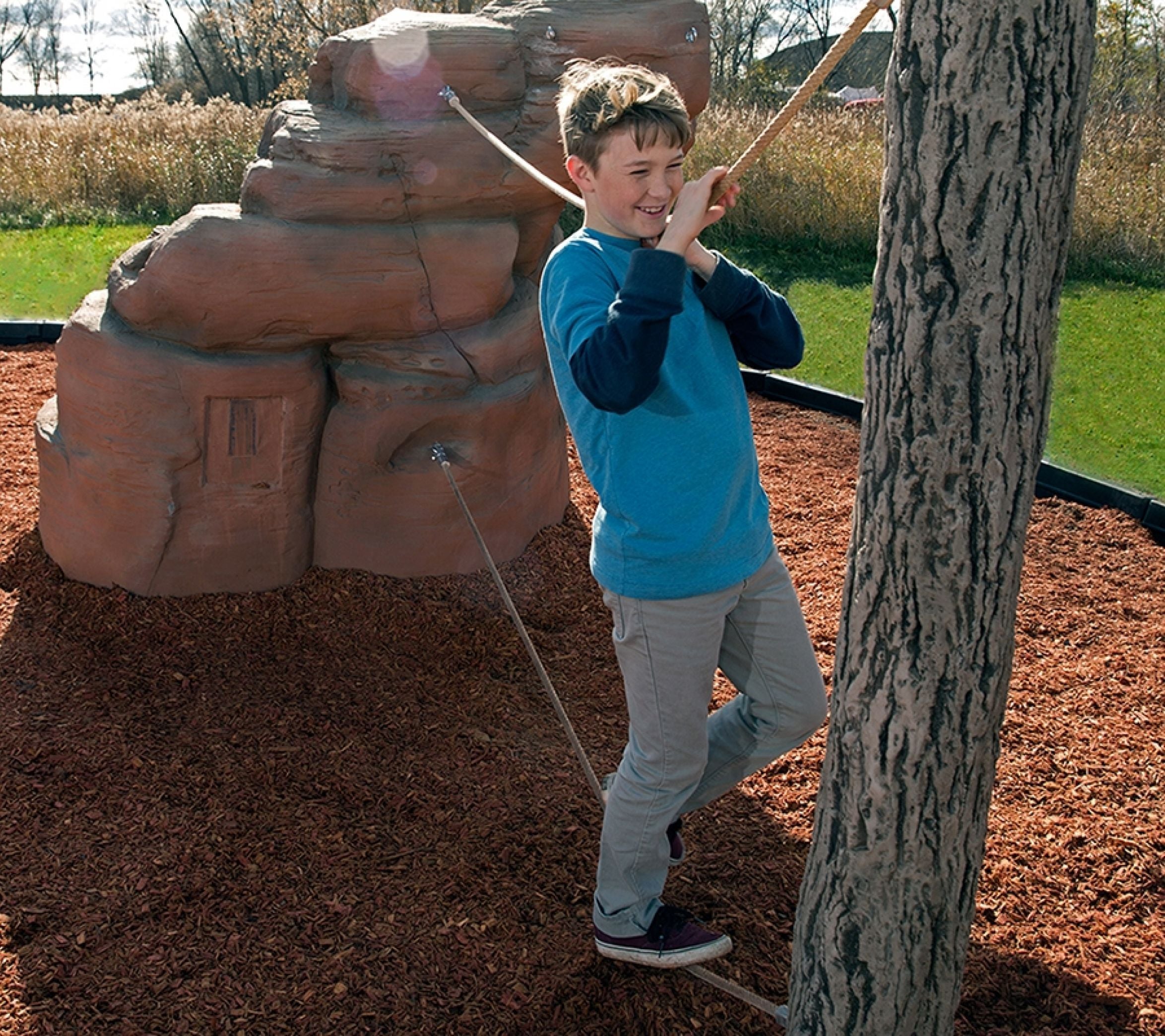 Child balancing on rope bridge between sandstone climbing boulder and textured log post in outdoor playground setting