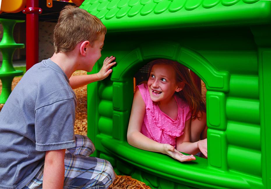Two children playing at a vibrant green playground panel shaped like a small house with a window opening.