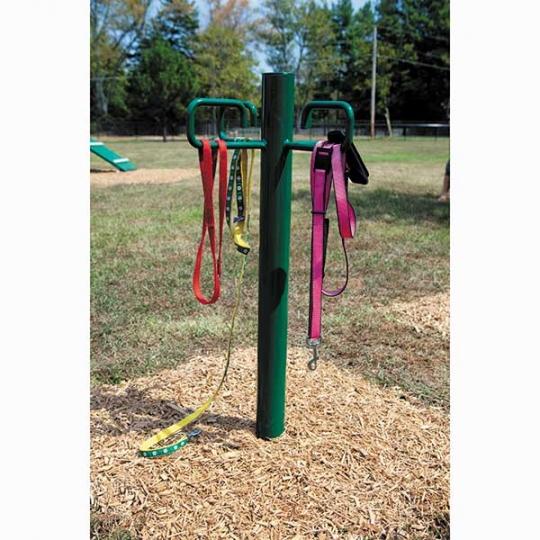 Green metal leash post with four C-hooks holding colorful dog leashes on wood chip ground in a park setting