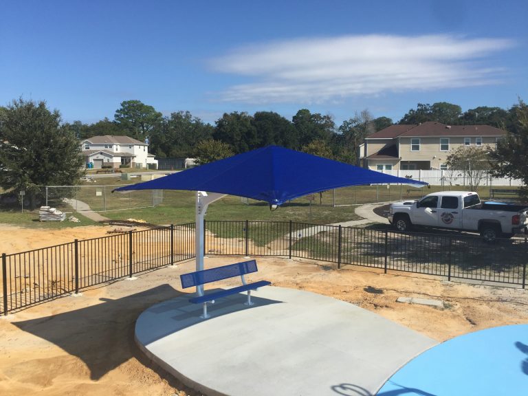 Blue cantilever square umbrella shade with white steel post shading a blue bench on a concrete pad in an outdoor park setting