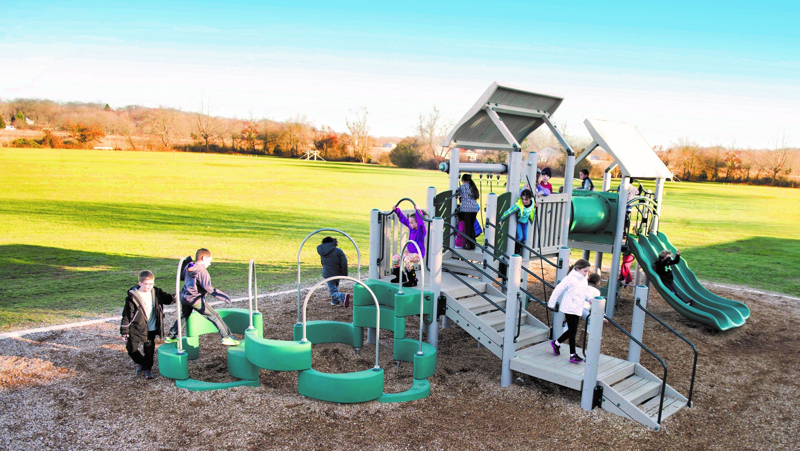 Outdoor accessible playground with climbing structures, tunnel crawl, triple wave slide, and wood chip surface with children playing