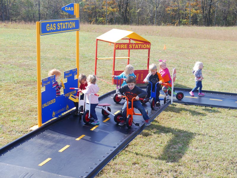 Children riding trikes on a winding outdoor play path with gas station and fire station play structures