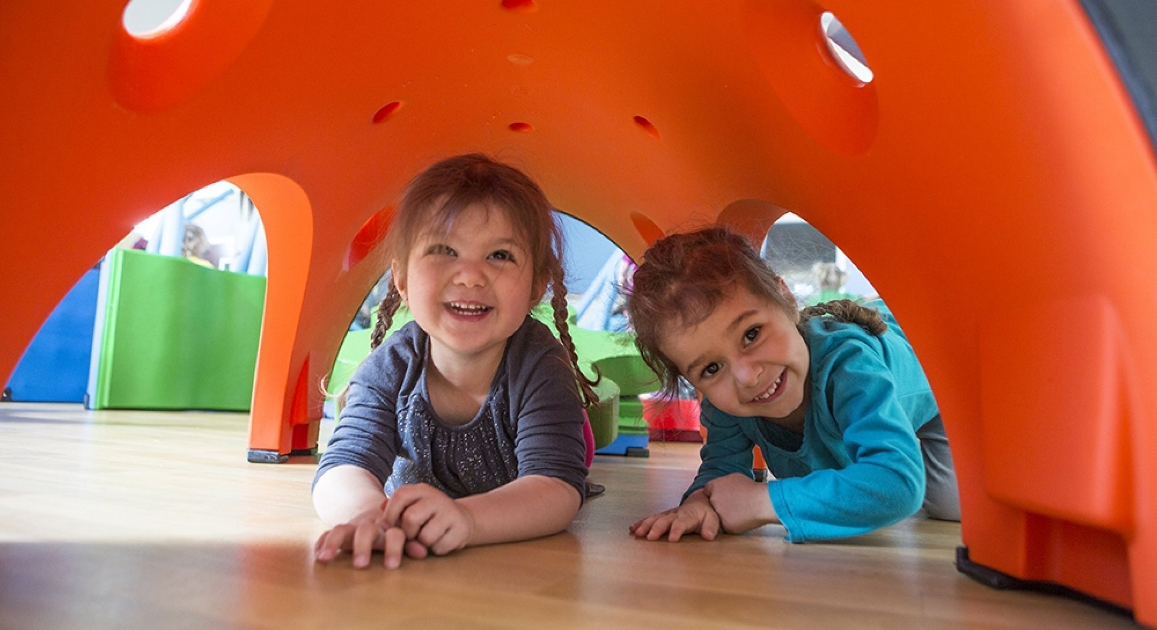 Two children crawling under an orange modular play structure in a colorful indoor play area designed for interactive, collaborative play