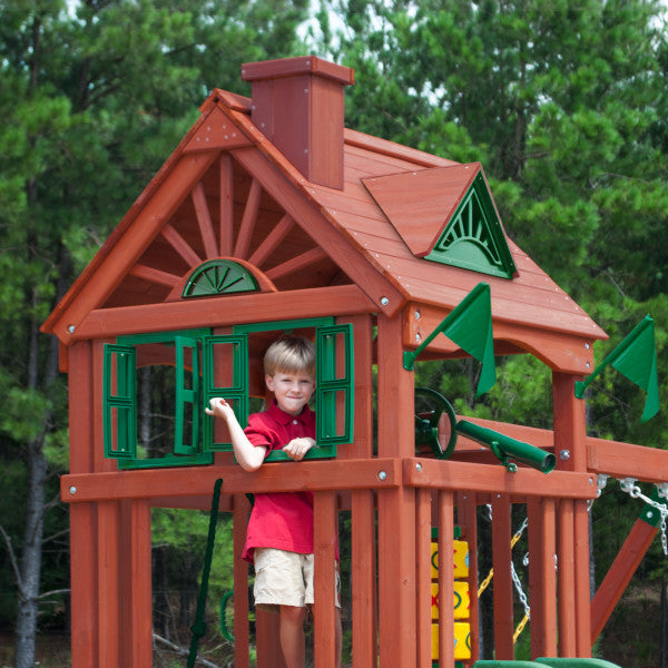 Cedar wooden playset with green shutters, two Alpine wave slides, rock wall, and child on deck surrounded by trees