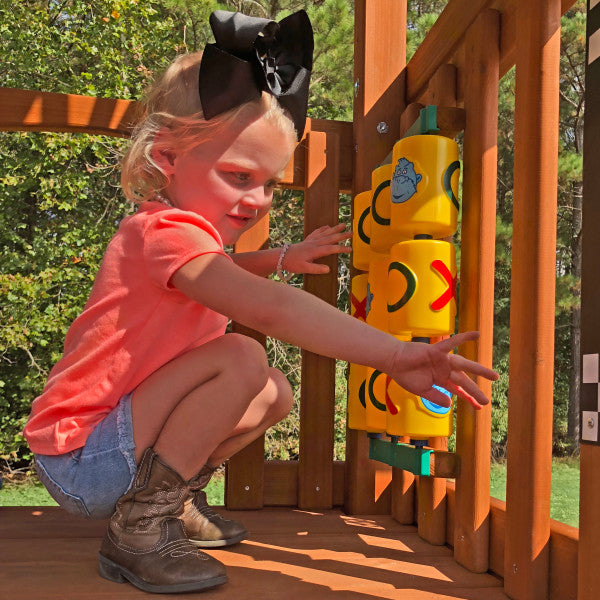 Young child with large black bow playing on wooden swing set deck with yellow tic-tac-toe panel surrounded by green trees