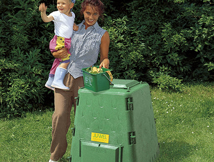Green durable outdoor compost bin with hinged lid, sliding lock, and two side doors, garden setting with woman holding child nearby