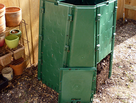 Green durable compost bin with vented side panels, hinged lid with sliding lock, placed outdoors near garden pots on soil ground