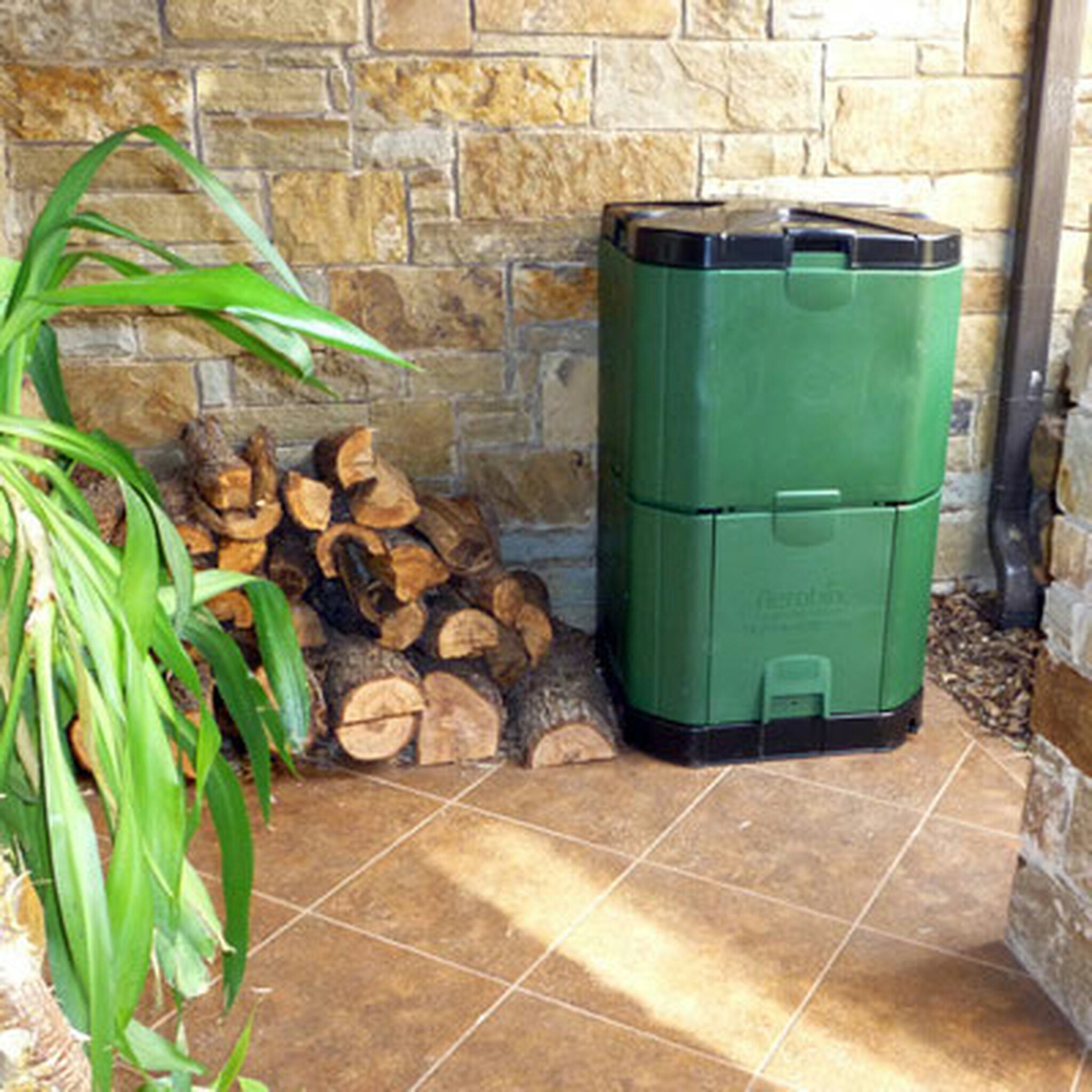 Green insulated compost bin with black lid and base, placed on tiled floor beside stacked firewood and a green plant
