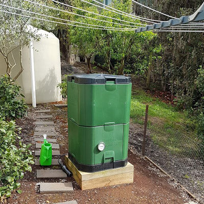 Green insulated compost bin with aeration core and leachate reservoir on wooden base near garden path