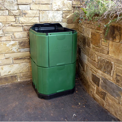 Green insulated dual-compartment compost bin with secure lid, placed in a stone-walled outdoor corner.