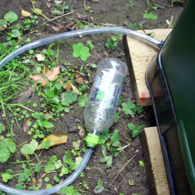 Green compost bin base with attached clear plastic tube and bottle on soil surrounded by leaves and wooden planks