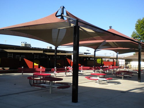 Modern outdoor shade structure with layered brown fabric panels supported by black metal poles over red picnic tables in a school courtyard