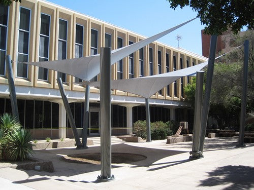 Hyperbolic fabric sail shade stretched between four tall metal columns in an outdoor courtyard setting.