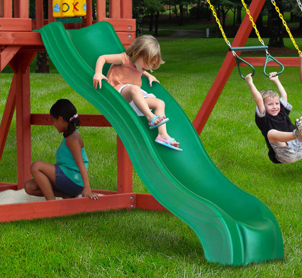 Cedar wooden playset on grass with green wave slide, two belt swings, trapeze bar, sandbox, and tic-tac-toe panel