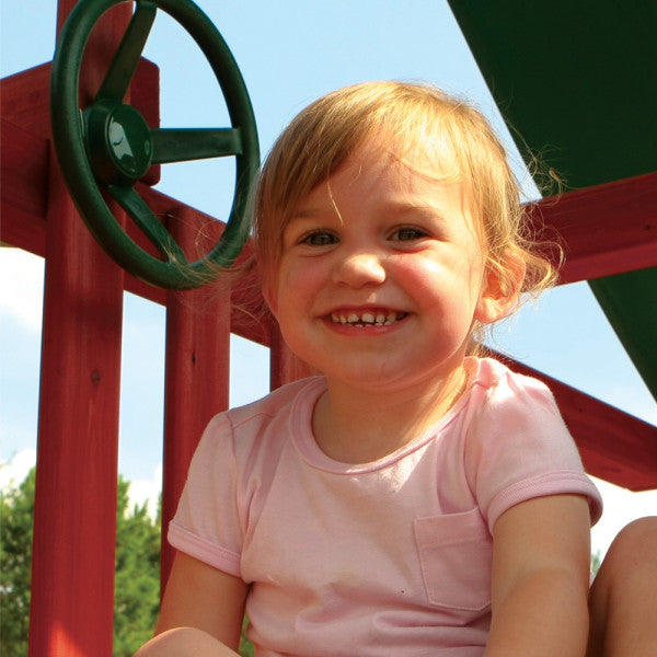 Toddler in pink shirt on redwood playset platform with green steering wheel and vinyl canopy above