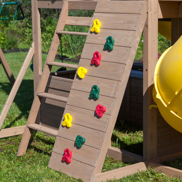 Wooden playset climbing wall with red, yellow, and green handholds beside yellow twist slide and ladder