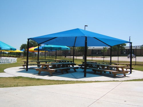 Blue hexagonal shade canopy with six black posts covering outdoor picnic tables in a park setting
