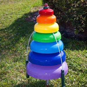 Seven rainbow-colored outdoor bell tree percussion bells with attached mallets on grass, designed for interactive musical play.