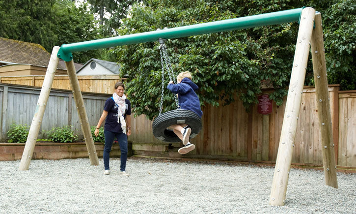 Child on a tire swing with green wooden frame in a gravel playground, promoting active play and coordination