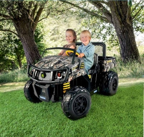 Two kids riding a black and camouflage John Deere Gator XUV toy vehicle with large wheels and rear cargo bed on grass