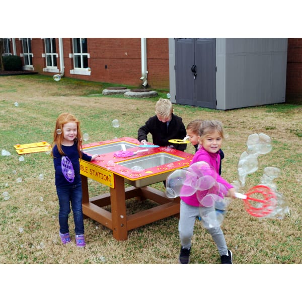 Children using an outdoor bubble station with wands and trays on grass near a building, promoting sensory and imaginative play