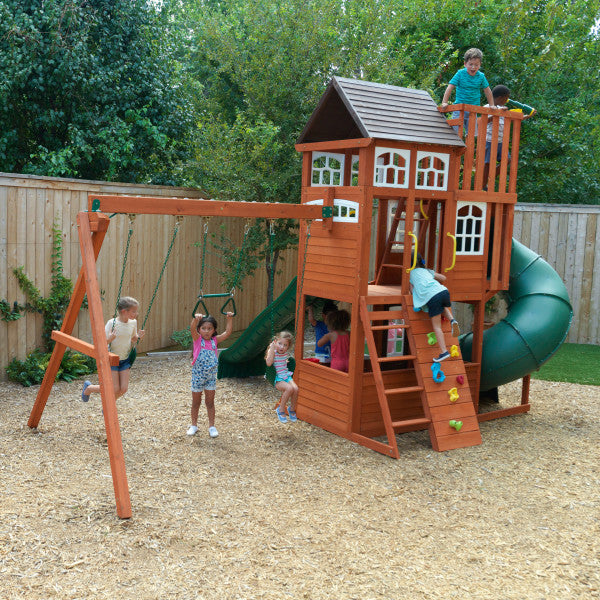 Wooden playset with climbing wall, tube slide, double wave slide, belt swings, acro bar, and kids playing on wood chip ground outdoors
